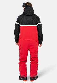 Chaqueta de nieve roja y negra con capucha, con rayas blancas. La tela parece texturizada, con un ajuste holgado y puños elásticos. Se ven gafas de esquí negras.