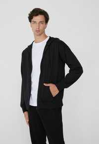 Felpa nera con zip realizzata in tessuto morbido, dotata di cappuccio con cordino, tasche frontali e polsini a costine. Indossata sopra una t-shirt bianca.