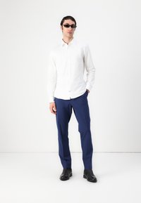 Chemise blanche à boutons associée à un pantalon bleu marine et des bottes en cuir noir. La chemise présente un col classique et une coupe ajustée.