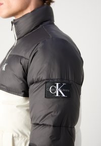 Melnbalts Calvin Klein pūkains mētelis ar augstu apkakli, kuram ir zīmola ielāps uz piedurknes un gluda, spīdīga auduma tekstūra.