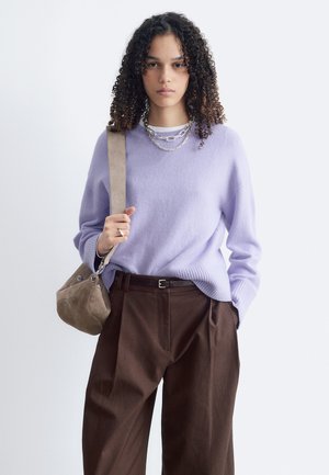 Jeune femme aux cheveux bouclés porte un pull violet clair, un pantalon marron, des colliers en argent et porte un sac à bandoulière beige sur fond blanc.