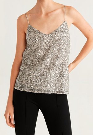 Femme portant un haut à fines bretelles argenté orné de paillettes et un pantalon noir ajusté, posant avec une main sur la hanche.