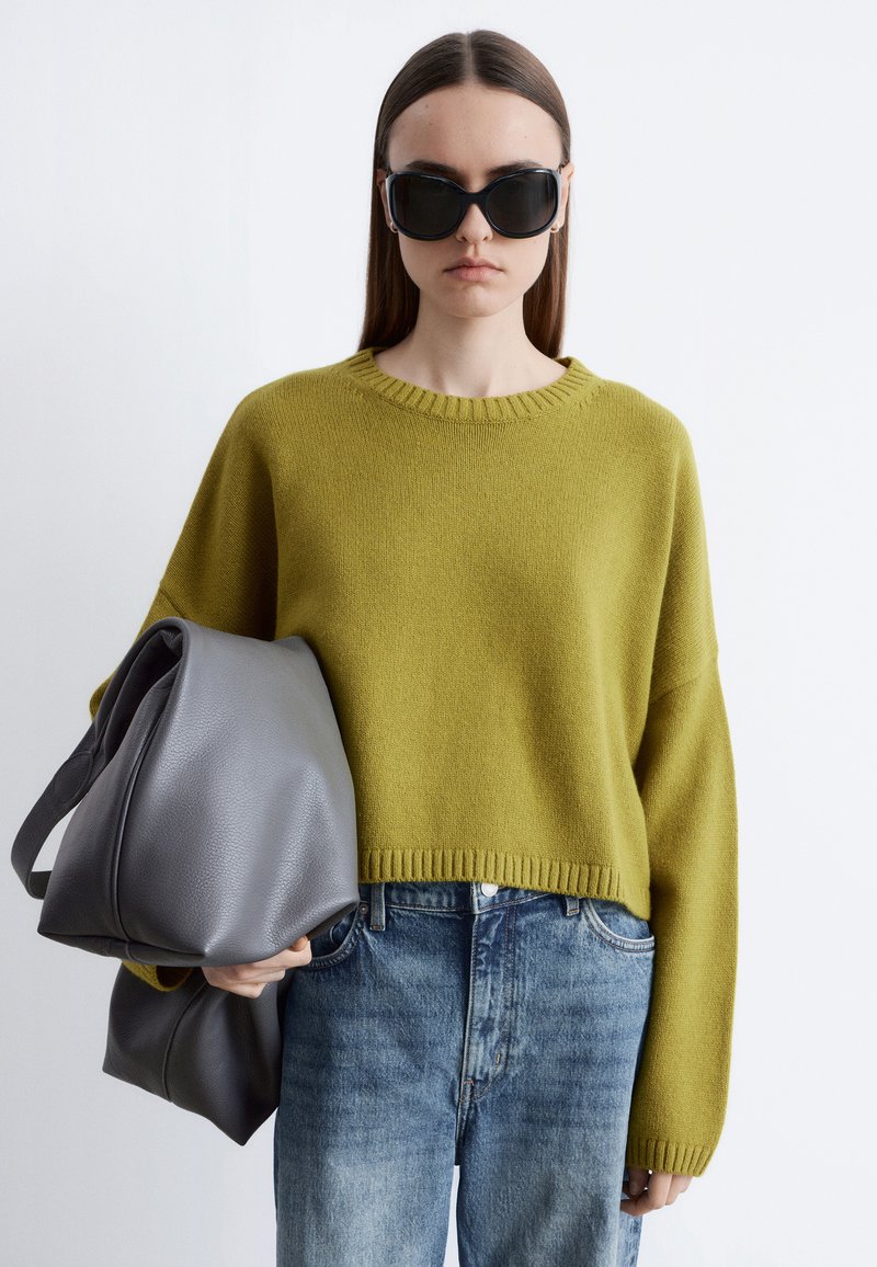 Mujer con gafas de sol negras de gran tamaño, suéter amarillo mostaza, vaqueros azules, sosteniendo un bolso grande de cuero gris sobre un fondo liso.