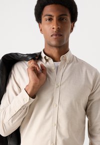 Μπεζ πουκάμισο με κουμπιά και χαλαρή εφαρμογή, έχει γιακά και μακριά μανίκια, με λεία υφή. Φοριέται με μια μαύρη δερμάτινη τζάκετ ριγμένη στον ώμο.