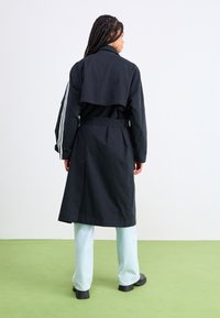Manteau noir avec une taille cintrée, un détail de rabat à l'arrière et trois rayures blanches sur les manches, associé à un pantalon bleu clair et des chaussures noires.
