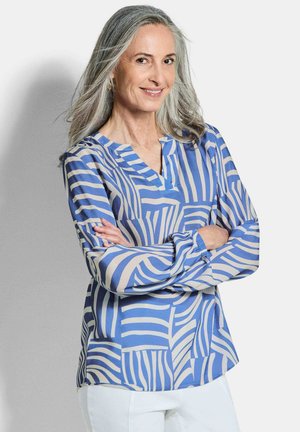 Reife Frau mit langen grauen Haaren, die eine blau-weiß gemusterte Bluse und weiße Hosen trägt, steht mit verschränkten Armen und lächelt.