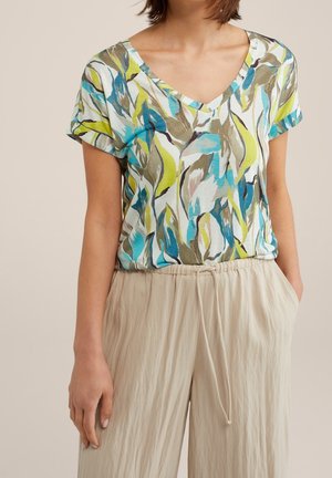 Kobieta w krótkim topie z dekoltem w serek z niebiesko-zielono-beżowymi wzorami liści, połączonym z beżowymi spodniami z troczkami.