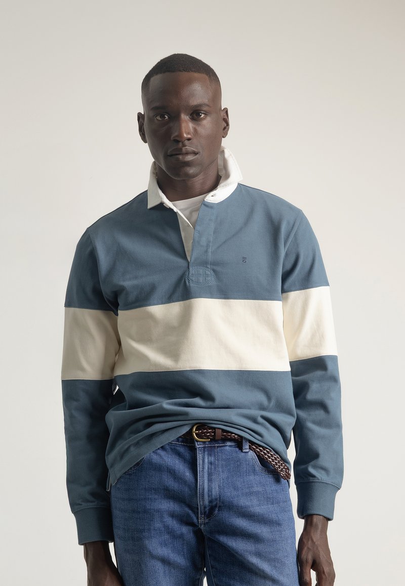 Polo de manga larga a rayas azul y crema con cuello, que presenta una tapeta de tres botones y puños en contraste. Llevado con jeans de mezclilla oscura.