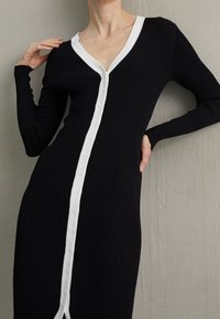 Donna che indossa un vestito nero aderente e a coste con rifiniture bianche sui bottoni e scollatura a V, mentre posano con una mano vicino al collo e l'altra sul fianco.