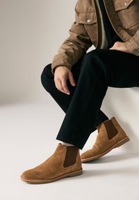 Bruine suède Chelsea-laarzen met elastische zijkanten, een afgeronde neus en een lichtbruine zool. Gecombineerd met een donkere broek en een bruine jas.