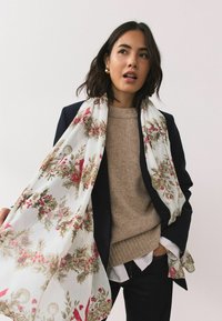 Šátek s květinovým motivem z průsvitné bílé látky, ozdobený červenými a zelenými botanickými detaily. Nošený přes béžový svetr a tmavé sako.