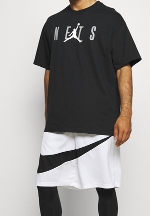 Osoba nosząca czarną koszulkę Nets z białym logo Jumpman oraz białe szorty z dużym czarnym logo Nike swoosh.