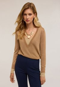 Haut à manches longues doré brillant avec un décolleté en V profond et des poignets beige contrastants, associé à un pantalon ajusté bleu marine et un collier épais crème.