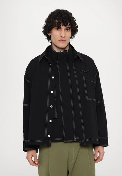 Black oversized shirt with white stitching details, featuring a button-up front, large collar, and a chest pocket, layered over a black top.