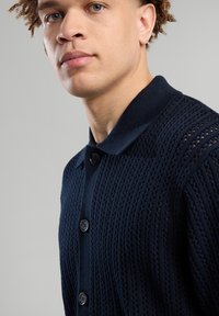 Laivaston sininen neulepusero, jossa on teksturoitu kuvio, polo-kaulus ja etupainatus, esittelee kevyttä materiaalia ja hengittävää muotoilua.