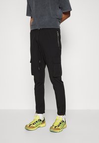 Svarta cargobyxor med justerbar dragsko i midjan, sidoficka med dragkedja, smal passform, kombinerade med gula och gröna sportskor.