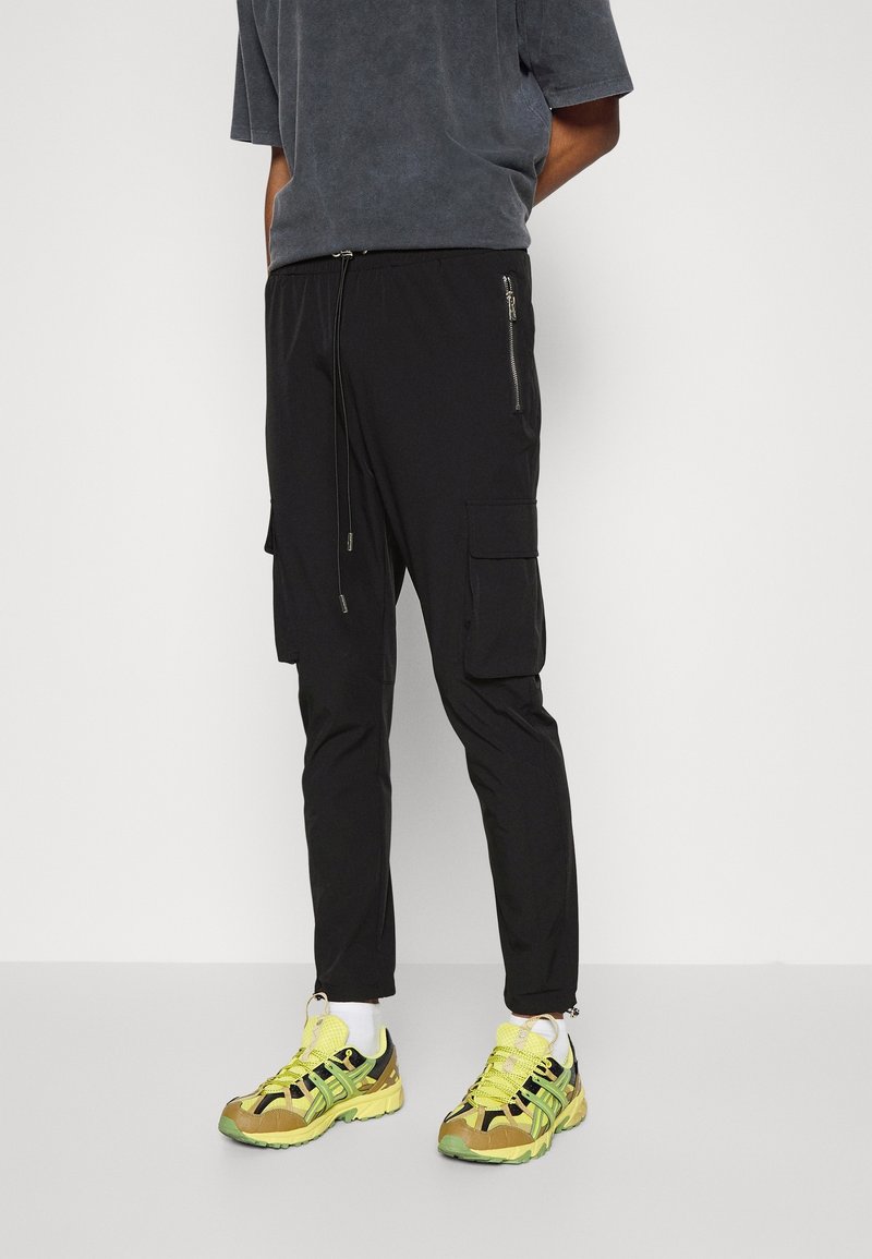 Svarta cargobyxor med justerbar dragsko i midjan, sidoficka med dragkedja, smal passform, kombinerade med gula och gröna sportskor.