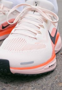 Svijetlopinkaste atletske cipele s mrežastim gornjim dijelom, naglašene narančastim detaljima i crnim Nike logotipom. Imaju vezivanje s čipkom i gumeni potplat.