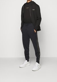Black zip-up hoodie, black t-shirt, and navy joggers with gold side logo. Completed with white sneakers. Soft, casual fit.
