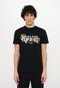 Juodas medvilninis marškinėliai trumpomis rankovėmis su baltu ir auksiniu "Versace Jeans Couture" logotipu ir dekoratyviniais gėlių elementais.