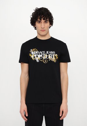 T-shirt en coton noir à manches courtes avec un logo « Versace Jeans Couture » blanc et doré ainsi que des éléments floraux décoratifs.