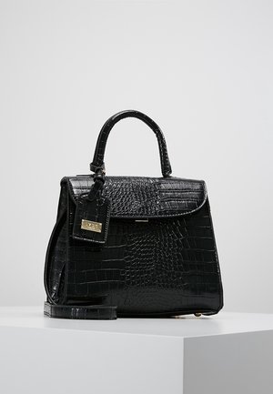 Czarna torebka ze skóry o fakturze krokodyla, z górnym uchwytem, klapką zamykającą, przymocowaną zawieszką oraz odpinanym paskiem na ramię, umieszczona na białym piedestale.