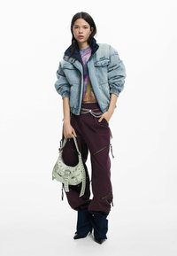 Geacă din denim cu guler bleumarin, top grafic colorat, pantaloni cargo mov cu fermoare și o geantă verde deschis cu ținte.