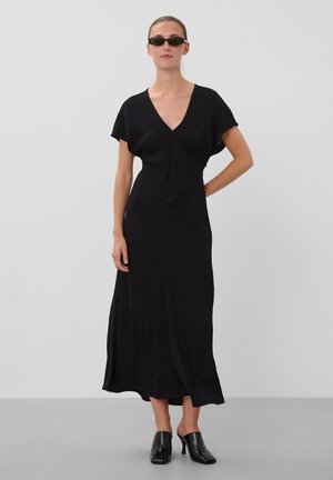 Femme portant une robe midi noire à col en V avec manches courtes, mules noires à talons et petites lunettes de soleil noires, debout devant un fond uni.