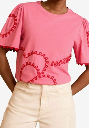 Rosa Kurzarmhemd mit roten, gestickten Schlaufenmustern an Ärmeln und Vorderseite, kombiniert mit hoch taillierten, cremefarbenen Hosen.