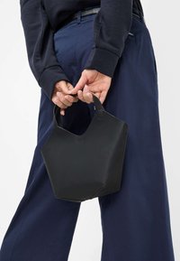 Zwarte leren tas met een geometrisch ontwerp, voorzien van korte handvatten, een gladde textuur en geen zichtbare hardware of patronen, vastgehouden door een persoon.