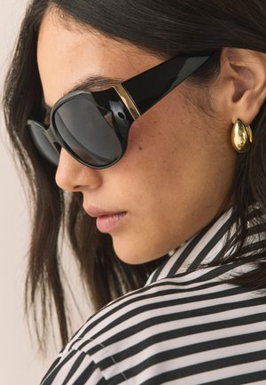 Mujer con cabello oscuro que lleva grandes gafas de sol negras, pendientes dorados en forma de lágrima y una camiseta de rayas blancas y negras, mirando hacia la izquierda en primer plano.