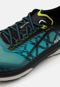 Scarpa sportiva con tomaia nera decorata da strisce ondulate in teal e aqua, motivi testurizzati, dettagli riflettenti e lacci a contrasto con particolare giallo neon.