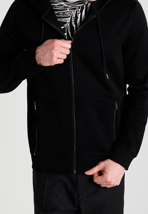 Person, die einen schwarzen Kapuzenpullover mit Reißverschluss trägt und diesen teilweise öffnet, um ein schwarz-weiß gemustertes Shirt darunter sichtbar zu machen.