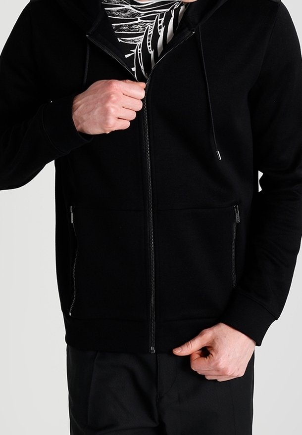 Personne portant un sweat à capuche noir zippé, en train de le dézipper partiellement pour révéler une chemise à motifs noirs et blancs en dessous.
