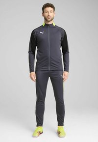 Mørkegrå atletisk træningsdragt med en jakke med lynlås, der har sorte ærmer og en neongul krave, sammen med matchende tætsiddende bukser og fodboldstøvler.