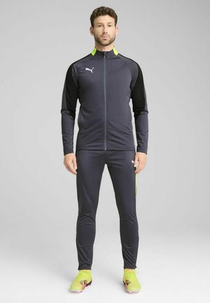 Dunkelgrauer Sportanzug mit einer Jacke mit Reißverschluss, die schwarze Ärmel und einen neonfarbenen gelben Kragen hat, kombiniert mit passenden schmal geschnittenen Hosen und Schuhen.