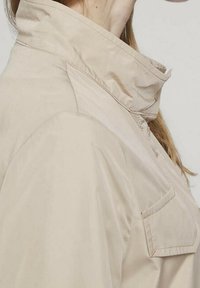Chaqueta beige con un cuello alto, bolsillo delantero y textura suave. Presenta un diseño simple y limpio, con un ajuste relajado.