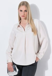 Blusa lunga manica beige chiaro con colletto, vestibilità ampia e dettagli plissettati sulla parte anteriore. Abbinata a una borsa a tracolla argento e pantaloni scuri.