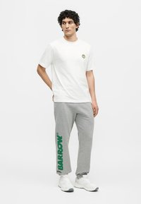 Jeune homme aux cheveux bouclés portant un t-shirt blanc avec un petit logo smiley jaune et un pantalon de survêtement gris avec le texte vert "BARROW" sur la jambe.
