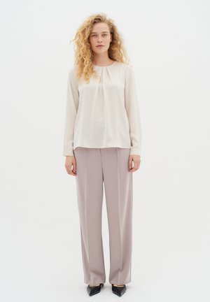 Donna con capelli biondi ricci, che indossa una blusa a maniche lunghe color crema, pantaloni a gamba larga rosa chiaro e scarpe nere a punta, in piedi su uno sfondo bianco.