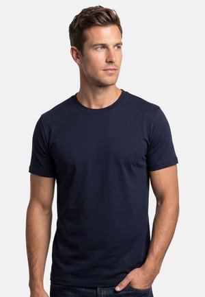Jonge man met kort bruin haar, gekleed in een effen donkerblauw T-shirt met korte mouwen, kijkt naar rechts met zijn hand in de zak.
