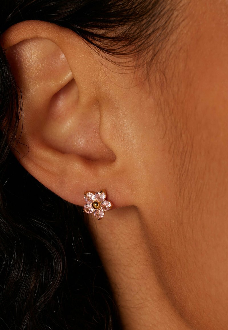 Gouden oorbel in de vorm van een bloem met vijf roze stenen en een centrale gouden kraal, geplaatst tegen een close-up van een oor. Gladde huidtextuur zichtbaar.