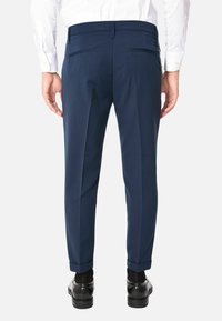 Pantaloni blu navy, slim fit, con tasche posteriori, texture liscia e orli arrotolati. Indossati con scarpe nere lucide.
