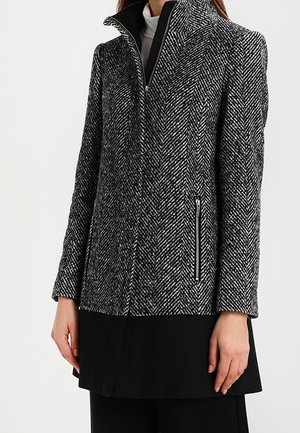 Vrouw draagt een grijs-zwarte jas met visgraatmotief en ritssluiting en hoge kraag, over een zwarte rok of jurk.