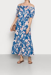 Robe longue dénudée sur les épaules en bleu avec un motif floral rose. Taille élastique, ourlet à volants, accompagnée de sandales métalliques à lanières et d'un petit sac.