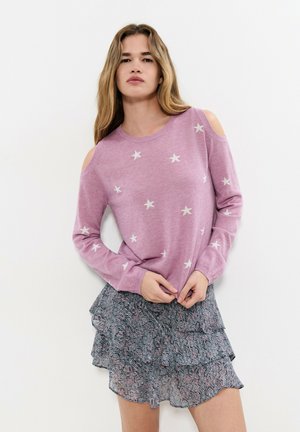 Mujer con un suéter rosa de hombros descubiertos con estrellas blancas y una falda de patrones en capas, sobre un fondo blanco liso.