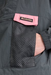 Veste noire avec une poche à rabat rose étiquetée Columbia Sportswear Company, avec une poche en mesh noire et une main dans une poche latérale.