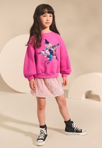 Roza široka majica z zvezdo iz modrih in roza bleščic, združena z roza krilom iz bleščic in črnimi visokimi supergami.