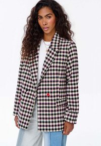 Žena s kudrnatými vlasy, oblečená v kostkovaném blejzru černé, bílé a růžové barvy, bílém topu, vrstvených náhrdelnících a dvoubarevných modrých džínách.