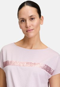 Jasnoróżowa bluzka z krótkim rękawem i szerokim dekoltem, ozdobiona poziomą taśmą z różnokolorowych cekinów na wysokości klatki piersiowej.